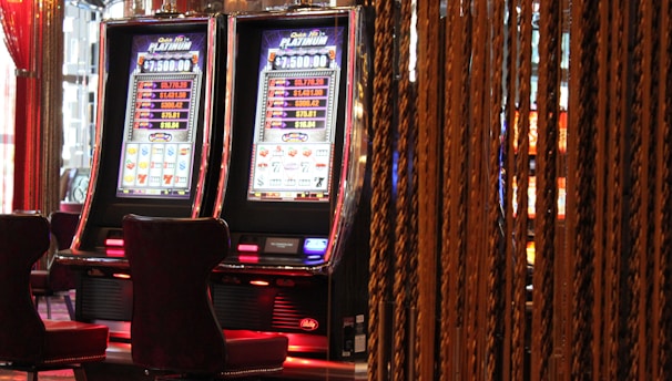 A pair of casino slot machines, each with detailed screens displaying various game outcomes and jackpot amounts. The chairs in front of the slot machines are dark and cushioned for comfort. The background features a hanging curtain made of golden ropes and other casino decorations are visible.
